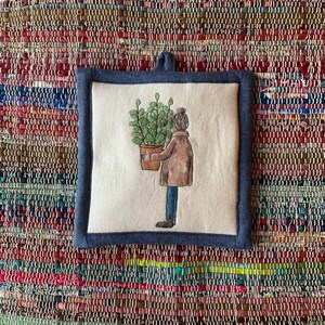 Może przedstawiać: Kwadratowy, kremowy podkładka pod garnek z tkaniny z akwarelową ilustracją osoby niosącej roślinę doniczkową. Podkładka ma niebieską dżinsową obwódkę i pętlę do zawieszenia. Tło to kolorowy tkany dywan.