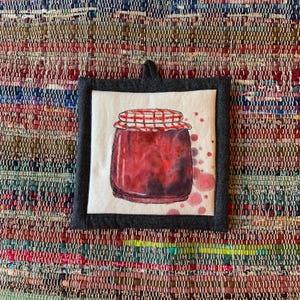 Peut inclure: Un dessous de plat carré en tissu avec une illustration à l'aquarelle d'un pot de confiture. Le pot est rouge avec un couvercle à carreaux blancs et rouges. Le dessous de plat a une bordure grise foncée et une boucle de suspension.