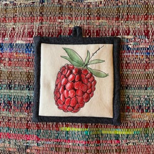 Peut inclure: Un dessous de plat carré de couleur écrue avec une illustration de framboise dessinée à la main. La framboise est rouge avec des feuilles vertes et une tige. Le dessous de plat a une bordure gris foncé et une boucle pour suspendre.