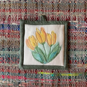 May include: Square, off-white pot holder with a watercolor painting of three yellow tulips with green leaves. The pot holder has a green border and a small loop for hanging. The background is a colorful woven rug.