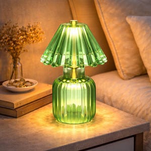 May include: A green glass table lamp with a ribbed base and a pleated lampshade. The lamp is lit, emitting a warm glow. The lamp sits on a light-coloured surface, beside a vase of dried flowers.
