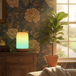 May include: A colour-changing LED lamp with a wooden base, displaying a gradient of yellow, green, and blue light. The lamp is on a wooden cabinet in a room with floral wallpaper and a potted plant. A window with natural light is in the background.