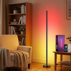 May include: A black LED corner floor lamp with colour-changing capabilities. The lamp is next to a small wooden table with a remote control and a smartphone. The lamp is in a living room setting with a bookshelf and armchair.