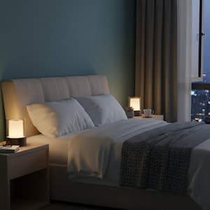 May include: A bedroom scene with a bed dressed in white bedding and pillows. Two bedside tables each hold a small, illuminated lamp. One lamp is cylindrical, the other is square. A window reveals a cityscape at dusk.