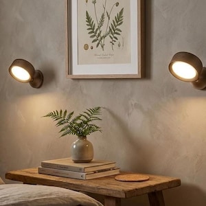 May include: Two wall-mounted, brown, adjustable spotlights illuminate a neutral-toned wall. A framed botanical print and a small wooden side table with a vase of greenery and books complete the scene. The warm lighting creates a cozy atmosphere.