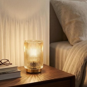 May include: A cylindrical, amber-colored glass table lamp with vertical ribbed detailing. The lamp sits on a gold-toned base and emits a warm, inviting glow. The lamp is on a wooden nightstand next to a stack of books and eyeglasses.