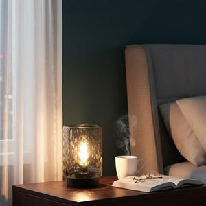 May include: A decorative table lamp with a textured glass shade, emitting a warm glow. The lamp sits on a dark wooden bedside table next to a white mug, an open book, and spectacles. A bed with white pillows is in the background.
