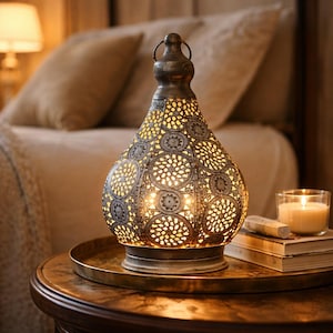 Op de afbeelding: Een decoratieve metalen lantaarn met een opengewerkt ontwerp, die een warme gloed uitstraalt. De lantaarn staat op een rond dienblad, naast een brandende kaars en boeken. De lamp heeft een handvat en staat op een houten tafel.