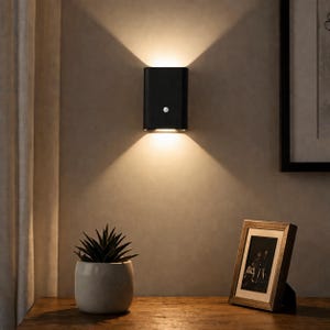 May include: A black wall-mounted light fixture with a sensor, casting light up and down. The sconce is on a neutral wall. A small potted plant and a framed photograph are visible below the light fixture.