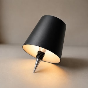 May include: A matte black bottle stopper featuring a conical shade design. The exterior is solid black, while the interior is white. It includes a silver-toned metal pointed tip and a black ribbed rubber seal for insertion into a bottle opening.