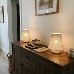 May include: Two table lamps with conical, off-white string-wrapped shades sit on a dark wood sideboard. Between them are a small wooden tray with keys, a white bowl containing dark objects, a notepad with a pen, and a small candle.