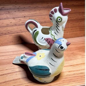 May include: Two ceramic bird-shaped containers. The back piece is a white rooster creamer with purple, green, and black painted details. The front piece is a white bird-shaped shaker featuring yellow, blue, and green floral and wing patterns.