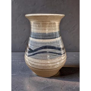 Può includere: Un vaso in ceramica con smalto color crema maculato caratterizzato da fasce orizzontali blu e grigie. Il vaso presenta un bordo svasato, un collo stretto e un corpo arrotondato con una finitura liscia e lucida che mette in risalto le sfumature.
