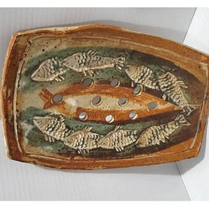 Könnte beinhalten: Eine rechteckige Seifenschale aus Keramik mit brauner und grüner Glasur. Die Oberfläche zeigt einen zentralen geprägten Fisch, umgeben von sechs kleineren Fischen im Relief. In der Mitte befinden sich sechs runde Abflusslöcher.
