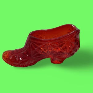 May include: A translucent red glass shoe figurine featuring a pressed diamond and starburst pattern across its exterior surface. The decorative object is shaped like a low-heeled pump with a hollow interior, showcasing a vibrant, deep red color.