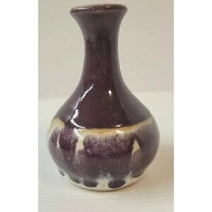 May include: A small, bulbous ceramic vase with a glossy finish. The body features a deep purple glaze that transitions into a creamy white drip pattern around the lower section. It has a narrow, cylindrical neck and a flared, rounded rim.
