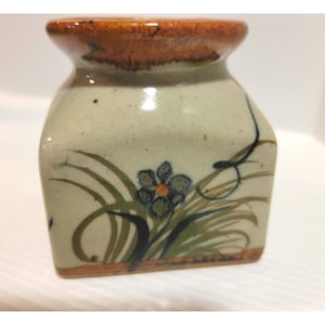 May include: A small, square ceramic vase with a light beige body and a dark brown rim. The vase is decorated with a floral design in shades of green, blue, and orange. The vase is approximately 4 inches tall.