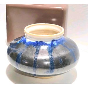 May include: A small, squat ceramic vase with a wide, rounded body and a narrow, circular opening. The exterior features a glossy, dark charcoal-grey base glaze with vibrant, cobalt-blue vertical drips cascading from the rim down the sides.