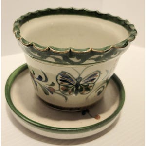 May include: Ceramic plant pot with a matching saucer. The off-white pot has a green scalloped rim and a painted butterfly design. The saucer has a green rim. The pot is suitable for indoor or outdoor use.
