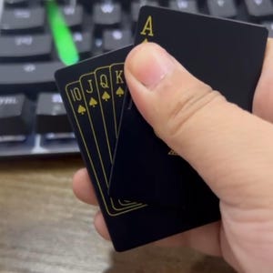 Puede incluir: Una mano sosteniendo una baraja de cartas negras con detalles dorados. Las cartas que se muestran son el 10, la J, la Q y la K de picas, junto con el As de picas. Las cartas tienen un diseño elegante y moderno.