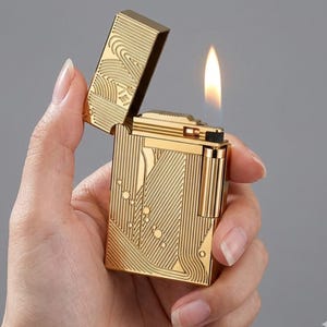May include: A gold-colored lighter with an open lid and a lit flame. The lighter has an engraved design on the lid and body. The design includes lines, circles, and wave-like patterns. The lighter is held in a hand.