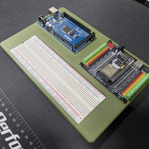 May include: An electronics prototyping kit featuring a blue ELEGOO Arduino board, a breadboard, and a black circuit board with various connectors. The components are mounted on a green base, ideal for electronics projects.