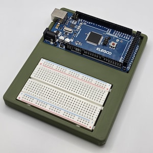 Peut inclure: Une carte Arduino ELEGOO avec un circuit imprimé bleu est placée dans un boîtier vert, avec deux plaques d'essai blanches en dessous. Les plaques d'essai ont des lignes rouges et bleues. Le boîtier est de forme rectangulaire.