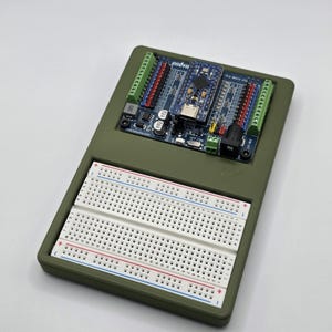 Puede incluir: Una carcasa electrónica verde oliva con una placa de circuito azul y una placa de pruebas blanca. La placa de circuito tiene múltiples conectores y componentes de colores. La placa de pruebas tiene filas de agujeros para conectar componentes electrónicos.