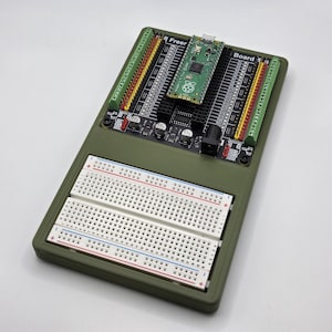 May include: An olive green electronics board with a Raspberry Pi Pico, breadboards, and various components. The board has multiple connectors and the word "Board" is visible. The word "Freer" is also visible.