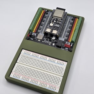 May include: An olive green electronics project board with a black circuit board, breadboard, and various connectors. The circuit board has multiple components and a small, silver module. The breadboard has red, blue, and black lines.