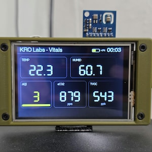 Könnte beinhalten: Ein olivgrünes elektronisches Gerät, das Umweltdaten anzeigt. Der Bildschirm zeigt Messwerte für Temperatur (22,3°C), Luftfeuchtigkeit (60,7), Luftqualitätsindex (3), eCO2 (879 ppm) und TVOC (543 ppb). Das Gerät ist mit "KRO Labs - Vitals" beschriftet.