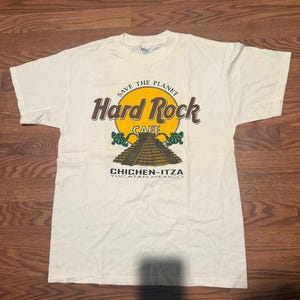 Könnte beinhalten: Cremefarbenes Hard Rock Cafe T-Shirt mit gelb-braunem Logo. Das Logo enthält die Wörter "Hard Rock Cafe" und "Chichen-Itza, Yucatan, Mexico". Das Shirt hat auch den Text "Save the Planet".