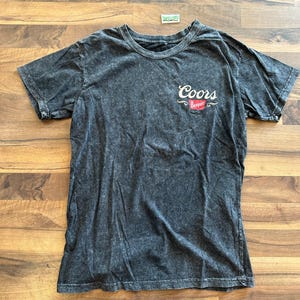 以下が含まれることがあります： クルーネックのダークグレーTシャツ。胸元に白と赤で「Coors Banquet」のロゴが入っています。生地は質感のあるウォッシュ加工が施されています。Tシャツは木製の表面に平らに置かれています。