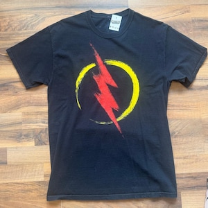 May include: Black t-shirt featuring a red lightning bolt design inside a yellow circle. The graphic is slightly distressed. The shirt has a crew neck and short sleeves. A small tag is visible near the neckline.