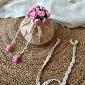 May include: A beige crocheted drawstring bag with a cluster of pink crocheted roses on top. The bag has a matching crocheted strap and two pink crocheted ball accents. The bag is sitting on a woven mat.