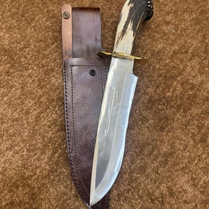 Peut inclure: Un couteau à lame fixe doté d'une lame en acier poli, d'une garde en laiton et d'un manche en bois de cerf texturé. Le couteau est accompagné d'un étui en cuir marron foncé avec des coutures visibles et une fermeture à pression métallique.