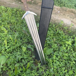 May include: A medieval-style sword with a wooden handle and a silver blade, resting in a black sheath. The sword's blade has a ridged design. The handle is a light brown color with small, round, gold-colored rivets. The sword and sheath are set in green grass.