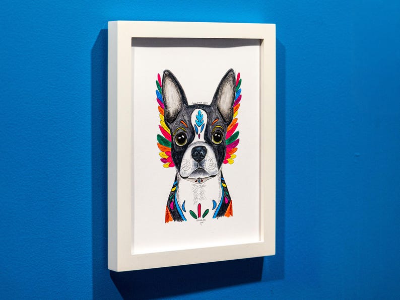May include: A framed art print featuring a Boston Terrier with colorful, stylized wings. The dog's face is black and white, with vibrant rainbow-colored accents. The artwork is in a white frame against a blue wall.