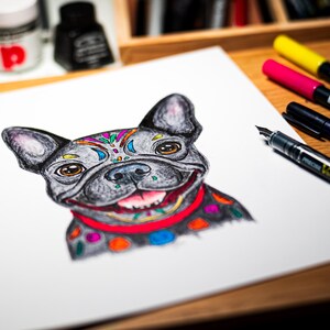 May include: A detailed drawing of a French Bulldog with colorful accents. The dog has a gray coat with decorative patterns on its head and collar. A red collar and a smiling expression complete the artwork. Pens and ink bottles are in the background.