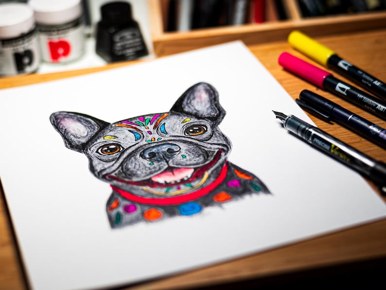May include: A detailed drawing of a French Bulldog with colorful accents. The dog has a gray coat with decorative patterns on its head and collar. A red collar and a smiling expression complete the artwork. Pens and ink bottles are in the background.