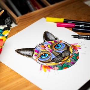 May include: A colorful cat illustration with blue eyes and floral accents. The artwork is on white paper, with markers and a pen nearby. The cat's face is decorated with various colors, including orange, yellow, and green.