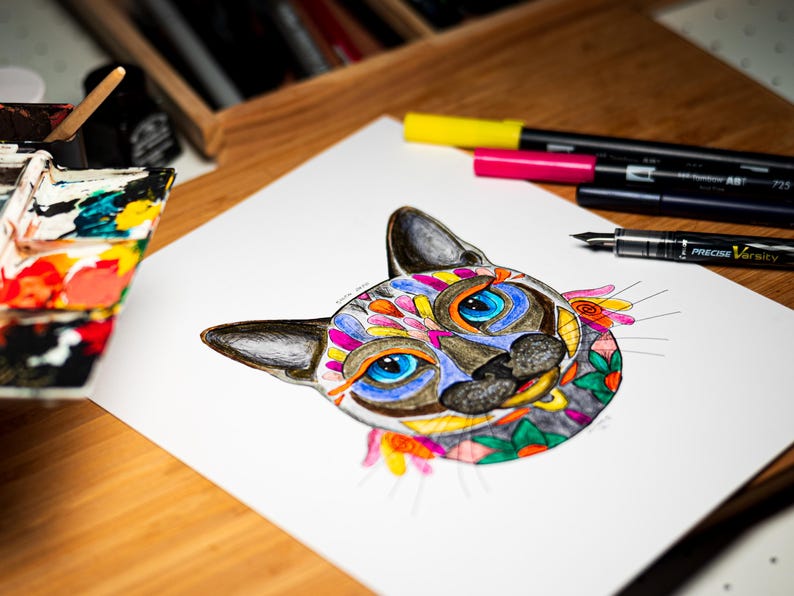 May include: A colorful cat illustration with blue eyes and floral accents. The artwork is on white paper, with markers and a pen nearby. The cat's face is decorated with various colors, including orange, yellow, and green.