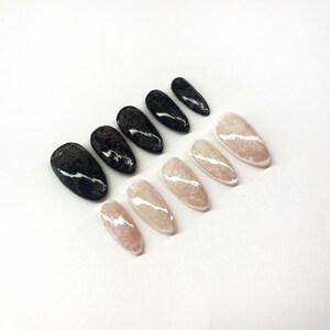 May include: A set of ten artificial nails in a variety of designs. Five nails feature a black lace pattern, while the other five have a nude base with a white, marbled design. The nails are arranged on a white surface.