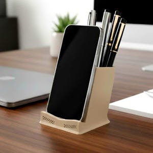 Może przedstawiać: Beżowy organizer biurkowy mieści smartfon i długopisy. Telefon jest czarny ze srebrną ramką. Długopisy są czarne i srebrne ze złotymi akcentami. Organizer stoi na ciemnym drewnianym biurku, z laptopem i rośliną w tle.