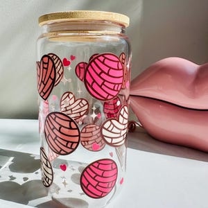 May include: A clear glass tumbler with a bamboo lid featuring a repeating pattern of pink and brown concha bread graphics, heart shapes, and small star icons printed on the exterior surface of the cylindrical glass container.