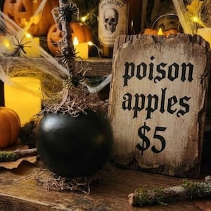 May include: A black round object with a twig stem and small plastic spiders, positioned next to a rectangular wooden sign with the text poison apples $5. The scene includes a brown glass bottle with a skull label and orange pumpkins.