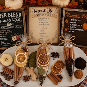 May include: Three clear cone-shaped bags filled with dried fruit, cinnamon sticks, and spices, tied with twine. Behind them are three wooden signs labeled Cider Blend, Harvest Blend, and Pumpkin Blend, listing ingredients like apples and coffee.