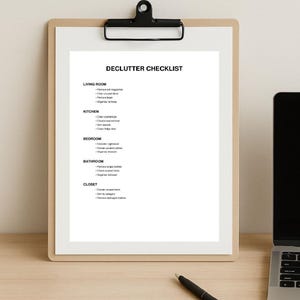 May include: A clipboard with a white paper checklist titled "Declutter Checklist". The checklist includes tasks for the living room, kitchen, bedroom, bathroom, and closet. A black pen and laptop are also visible.