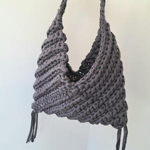 May include: A dark gray hobo-style bag crafted from thick, braided fabric yarn. The bag features a textured, crocheted pattern with a single integrated shoulder strap and two dangling fabric tassels at the bottom corners, secured with a metal snap.
