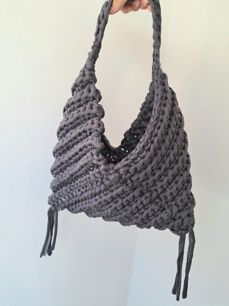 May include: A dark gray hobo-style bag crafted from thick, braided fabric yarn. The bag features a textured, crocheted pattern with a single integrated shoulder strap and two dangling fabric tassels at the bottom corners, secured with a metal snap.